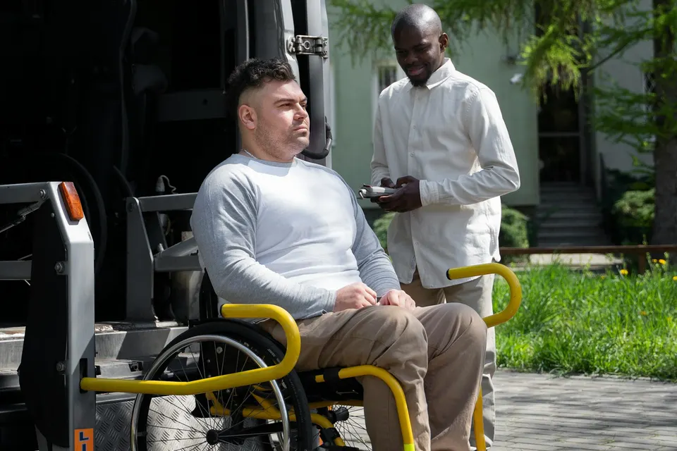 A man in a wheelchair on a van lift beside a driver holding a tablet