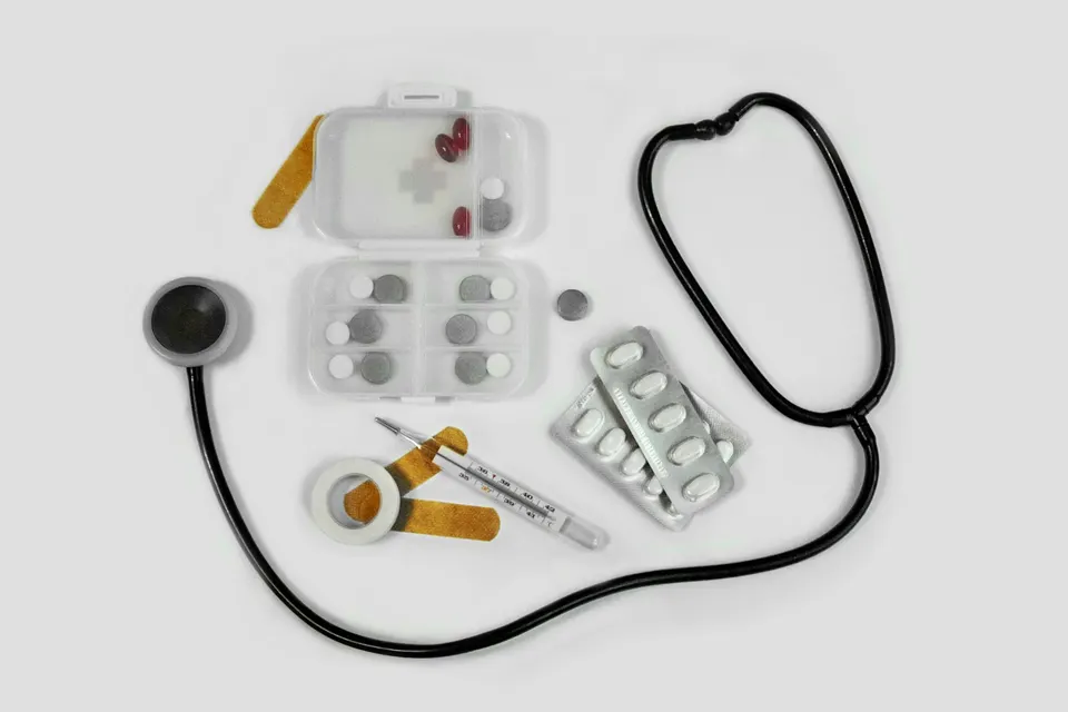 A stethoscope, pill organizer, thermometer, and bandages laid out on a white surface