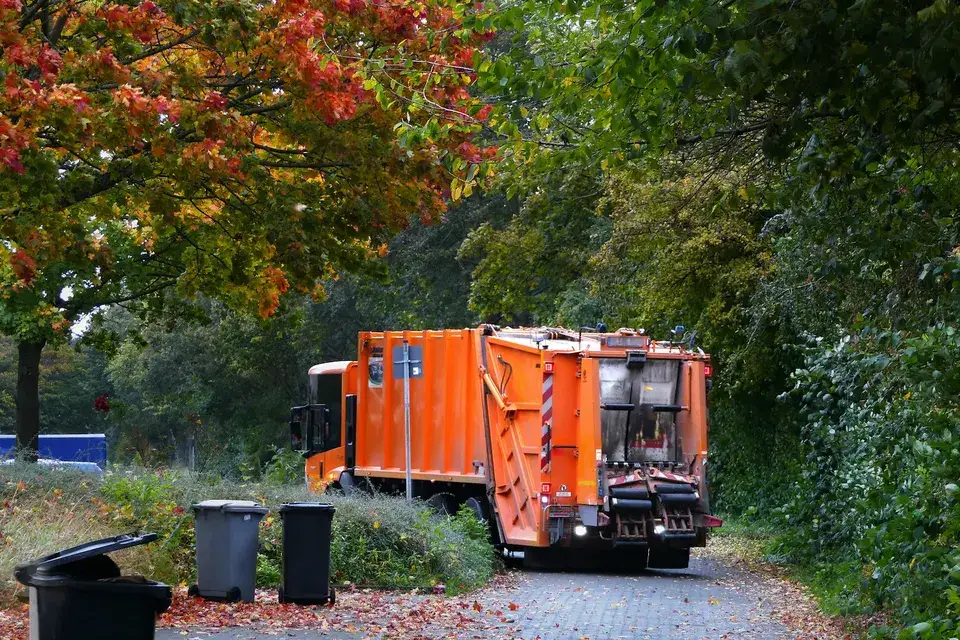 An orange refuse truck on a tree-lined lane in autumn, with bins at the curb